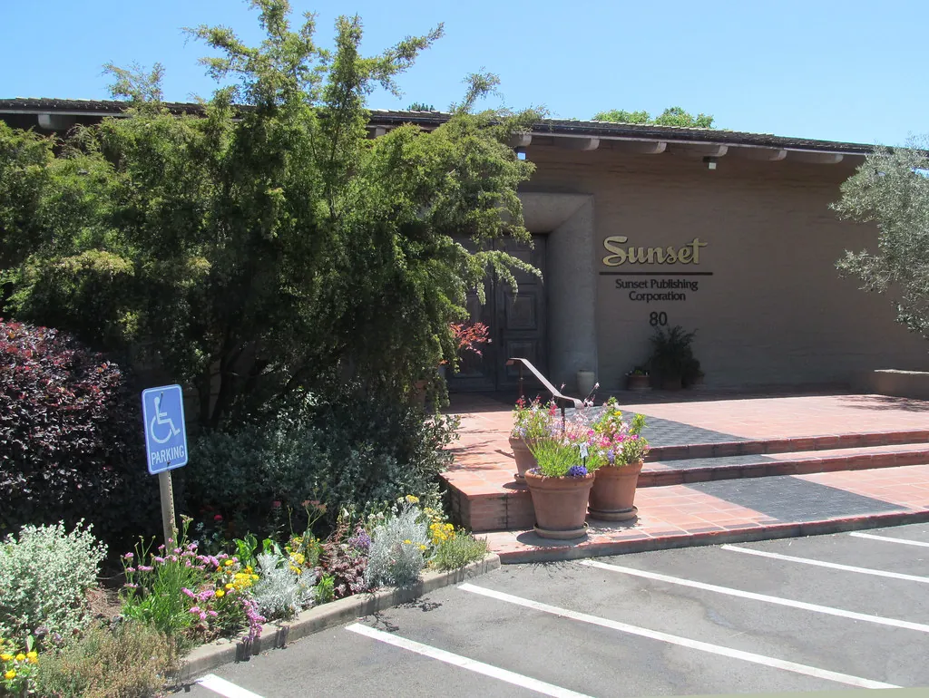 The entrance to the old Sunset Magazine headquarters: a low one-story building with the Sunset Publishing Corporation sign and the number 80, brick steps with potted flowers, and gardens beside the parking lot.